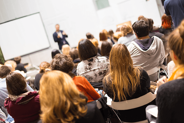 A seminar room full of people and a speaker standing at the front presenting and talking in a microphone.