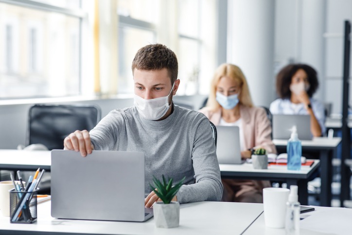 Coronavirus quarantine and office work with colleagues keeping social distancing. Portrait of young man in protective mask make video call with client at table with laptops with antiseptic