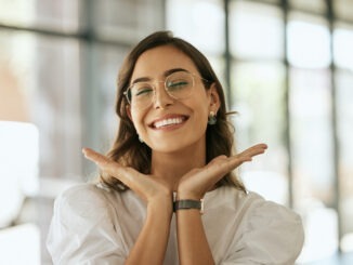 Cheerful business woman with glasses posing with her hands under her face showing her smile in an office. Playful hispanic female entrepreneur looking happy and excited at workplace